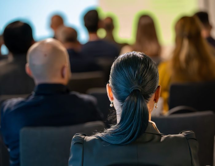 Eine Gruppe von Konferenzbesuchern, von hinten gesehen, mit Fokus auf eine Frau mit Pferdeschwanz
