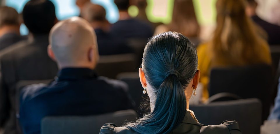 A group of people attending a conference, viewed from the back, focusing on a woman with a ponytail
