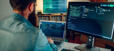 Rear view of a full-stack developer typing code on his laptop at Back view of a full-stack developer programming on a laptop at his home surrounded by two monitors. A senior developer is sitting at his home office and typing code on a laptop.