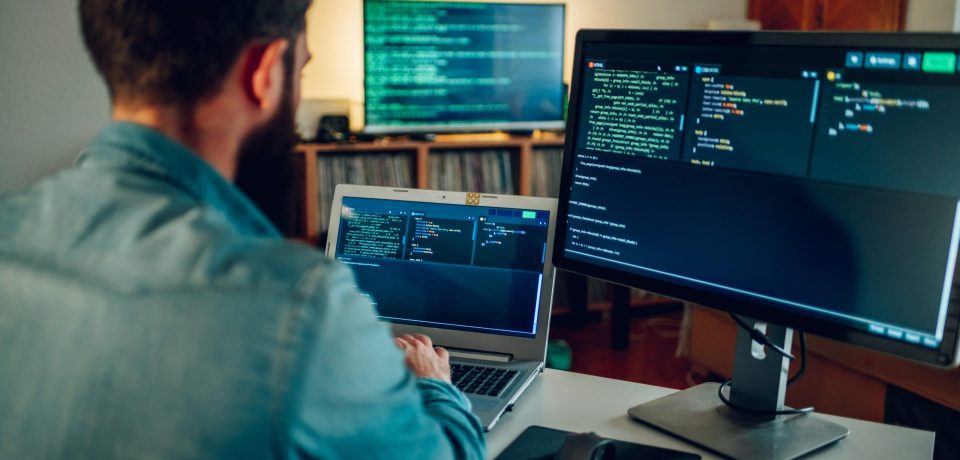 Back view of a full-stack developer programming on a laptop at his home surrounded by two monitors. A senior developer is sitting at his home office and typing code on a laptop.